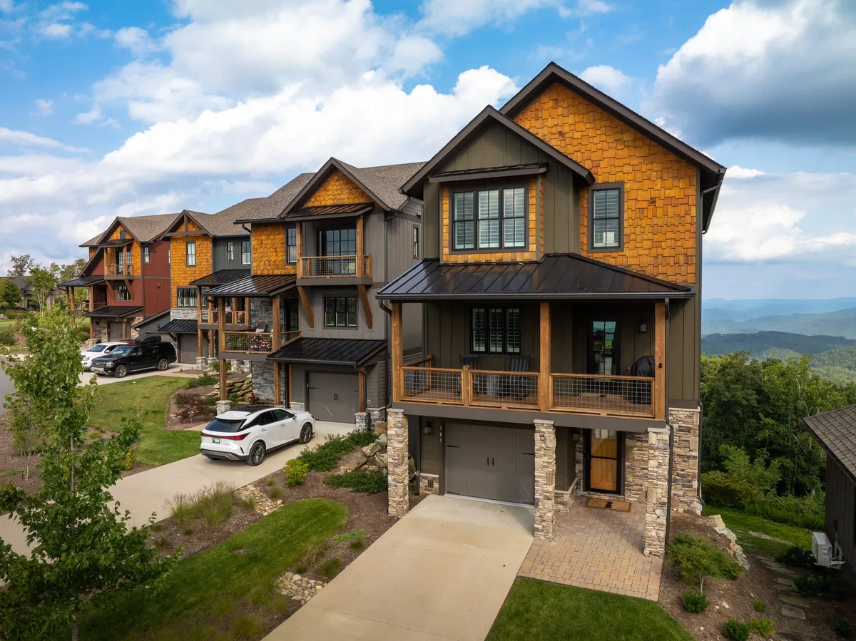 The image showcases a beautifully crafted multi-level mountain home with warm wooden accents and sturdy stone pillars, set amidst lush greenery and panoramic views of the Blue Ridge Mountains. Wide porches hint at inviting spaces perfect for savoring mountain sunsets, relaxing with family, or gathering for outdoor meals. The overall mood is welcoming and tranquil, ideal for those seeking both comfort and breathtaking scenery.