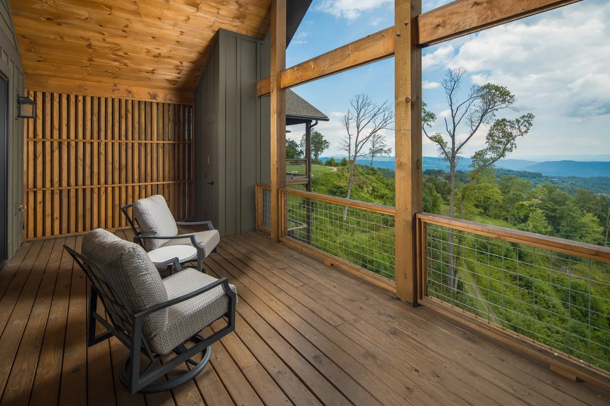 A serene porch with several rocking chairs facing a wooden railing, offering a panoramic mountain view. Sunlight filters through the trees onto the deck, evoking a peaceful, inviting atmosphere perfect for relaxing, reading, or simply savoring the fresh mountain air.