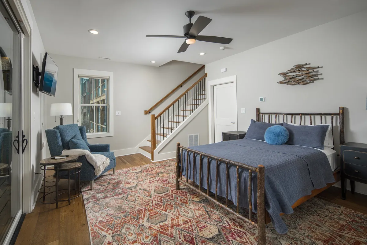 A queen bedroom featuring a rustic wooden frame bed, a patterned area rug, a cozy armchair and side table, and a staircase leading to another level. The room is bathed in natural light, highlighting its calm and inviting atmosphere.