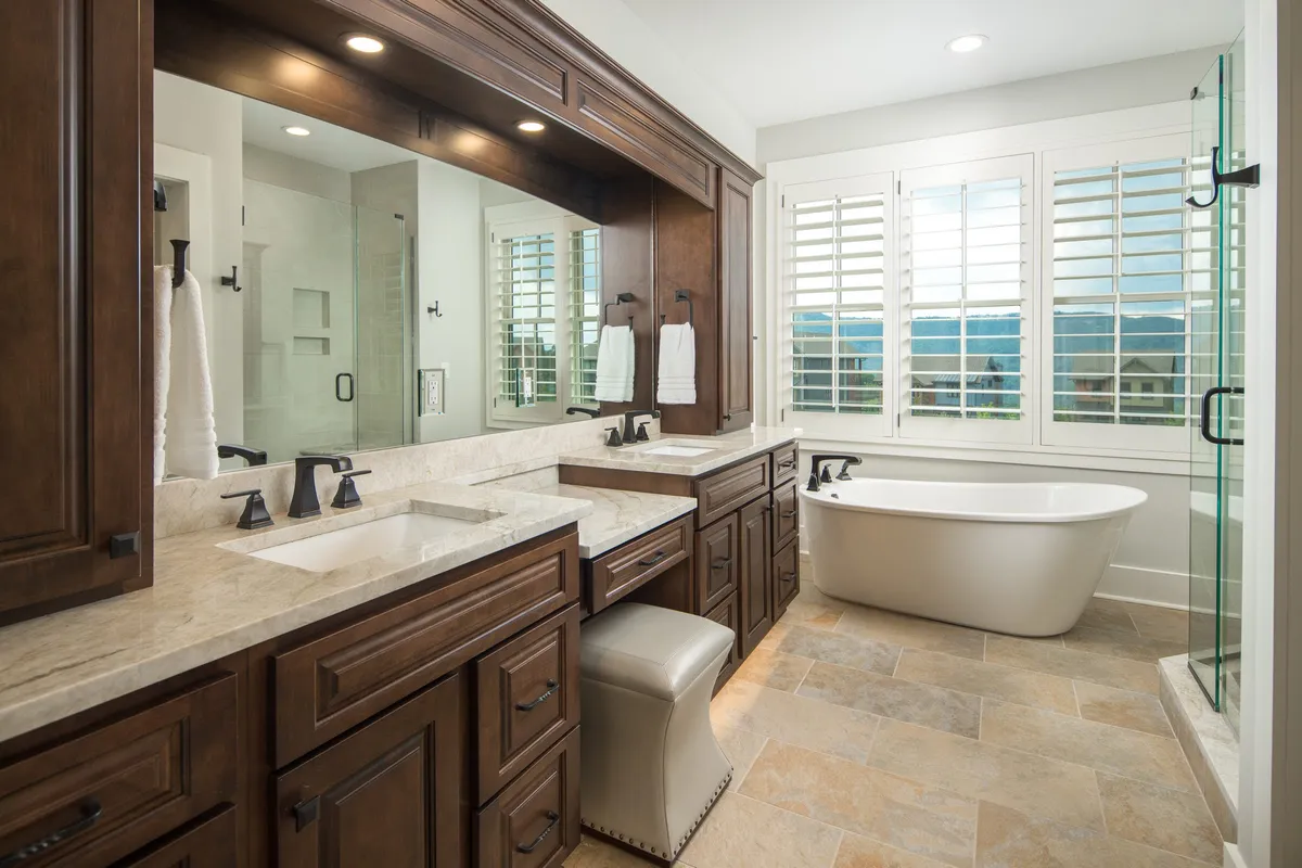 A luxurious bathroom with double sink vanities, a freestanding soaking tub, large mirrors, and shuttered windows allowing natural light to fill the space. The clean, airy design and spacious layout evoke a spa-like feel.