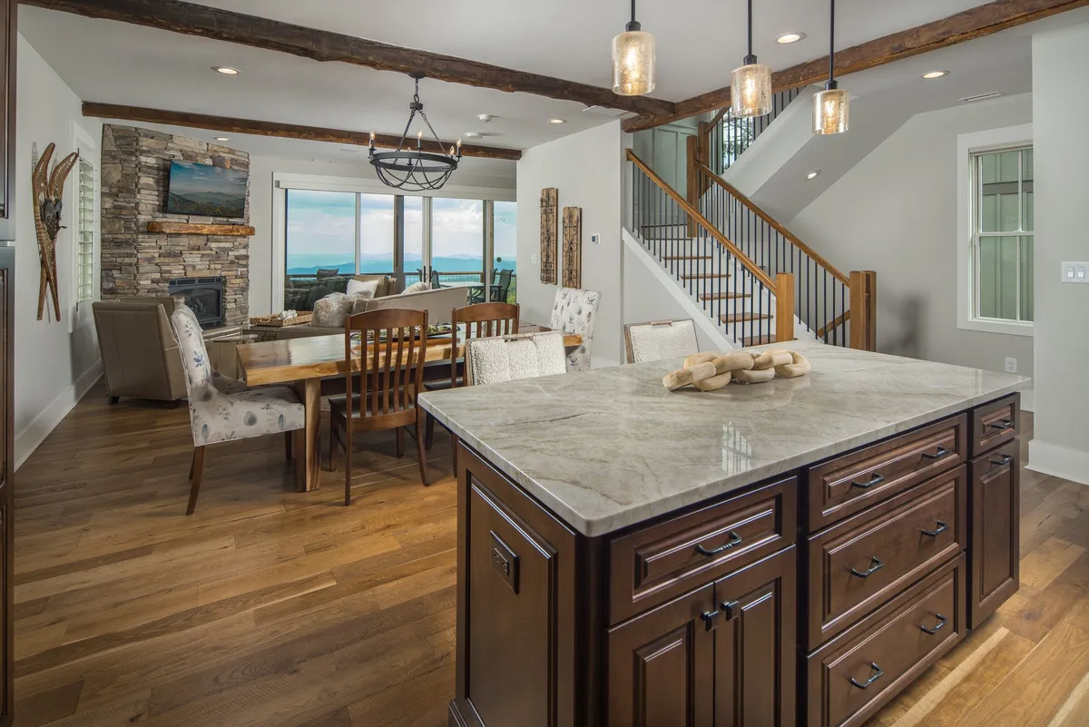 A wide view of the integrated kitchen, dining, and living spaces, with a large kitchen island, a family-sized dining table, and a fireplace. Expansive windows allow natural light to flood the room and offer picturesque views of the mountains beyond. The arrangement encourages interaction and shared moments.