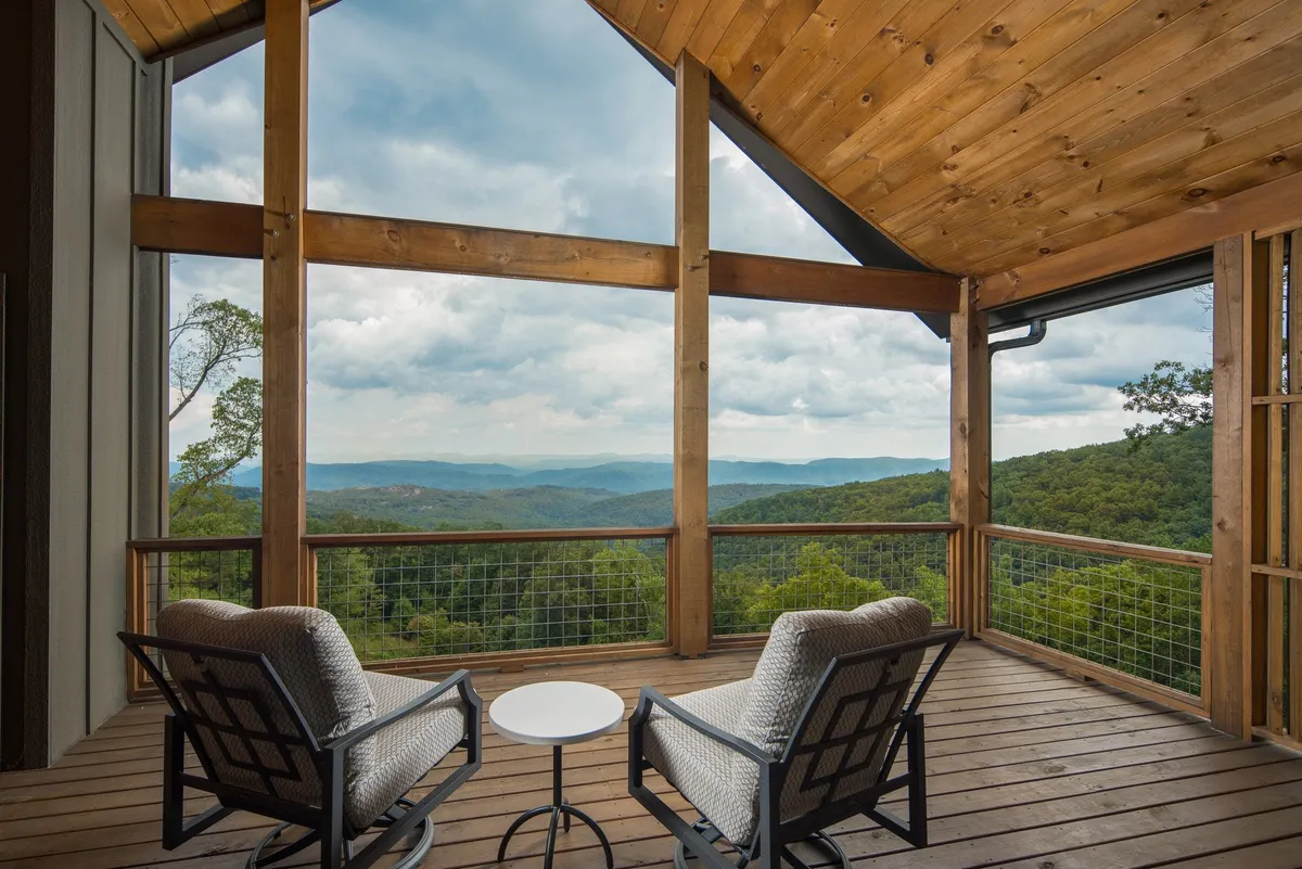 Two comfortable chairs sit on a small patio, separated by a round table perfect for drinks or morning coffee. Beyond, an expansive and unobstructed mountain view stretches into the distance, framed by natural greenery. The setting is tranquil, with no immediate distractions, inviting moments of stargazing or sunrise watching.