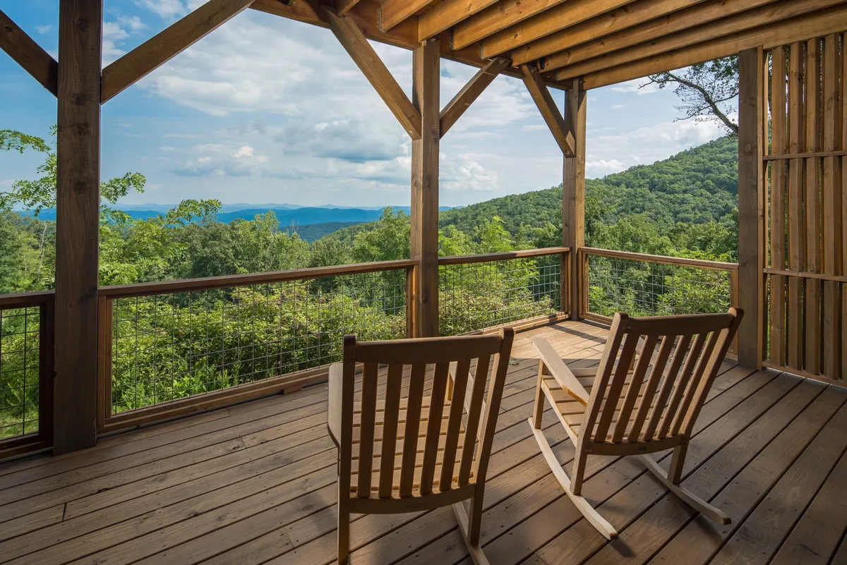 A pair of classic rocking chairs rest on a wooden deck, facing out toward a sweeping mountain landscape. Heavy wooden beams frame the space, creating a sheltered spot for relaxation. The view is peaceful and immersive, evoking a deep sense of connection with nature.