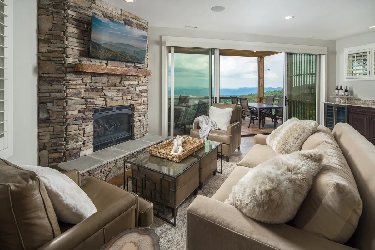 This image showcases the open living area, featuring plush sofas gathered around a striking stone gas fireplace. Large sliding glass doors frame sweeping mountain views, seamlessly connecting the living space to the outdoors. Decorative touches and a coffee table complete the warm, inviting atmosphere.