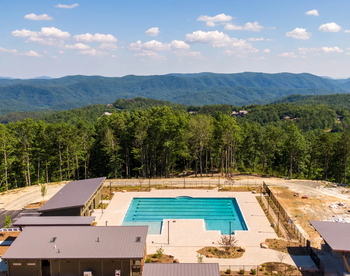 An inviting swimming pool sits at the edge of a landscaped area, with a stunning mountain backdrop and surrounding trees. The scene is sunlit and suggests both relaxation and recreation, with plenty of space for sunbathing, gentle swims, or simply gazing at the scenery.